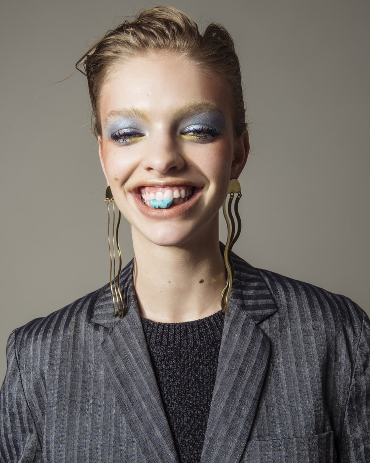Close-up of a smiling young woman with dramatic makeup wearing a grey pinstripe blazer and long gold earrings against a neutral background.