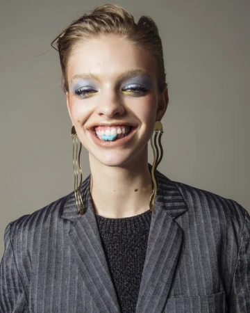 Close-up portrait of a smiling model wearing blue eyeshadow, long wavy gold earrings, and a pinstripe blazer by Matthew Priestley