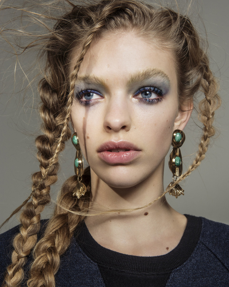 Beauty shot of a female model with blonde braided hair, dramatic blue eye makeup, and large turquoise and gold earrings. Studio lit.