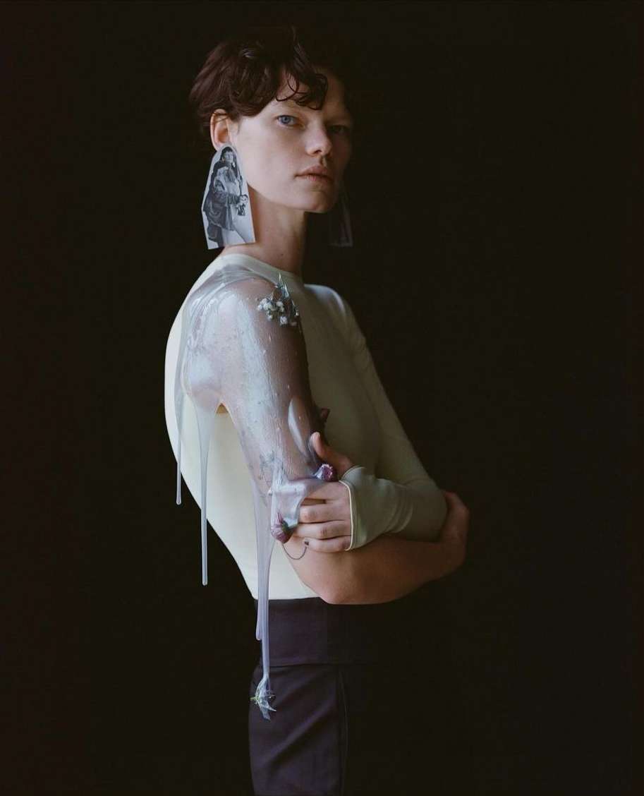 Woman in avant-garde fashion poses in a studio with artful makeup, sheer sleeves, and statement earrings against a dark backdrop.