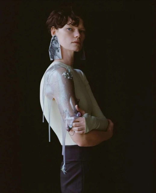 Woman in avant-garde fashion poses in a studio with artful makeup, sheer sleeves, and statement earrings against a dark backdrop.