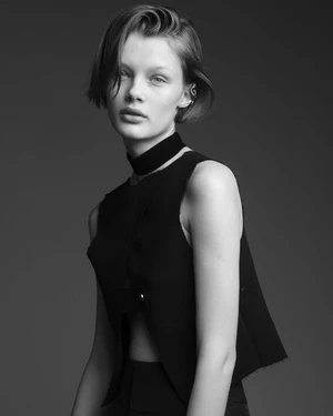 Black and white portrait of a fair-skinned young woman with short hair wearing a black vest and choker against a neutral gray backdrop.