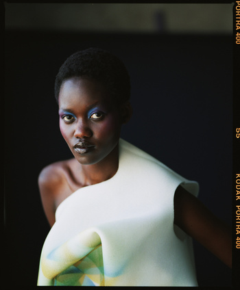 Portrait of a woman with blue eyeshadow and short hair wearing a sculptural white dress against a dark background in a studio.