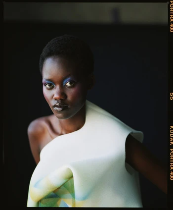 Portrait of a woman with blue eyeshadow and short hair wearing a sculptural white dress against a dark background in a studio.