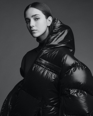 Portrait of a woman wearing a glossy black puffer jacket with a hood against a plain grey studio backdrop by Matthew Priestley.