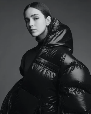 Portrait of a woman wearing a glossy black puffer jacket with a hood against a plain grey studio backdrop by Matthew Priestley.