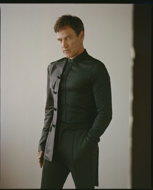 Man wearing a dark patterned button-down shirt and a half-leather jacket standing against a plain background