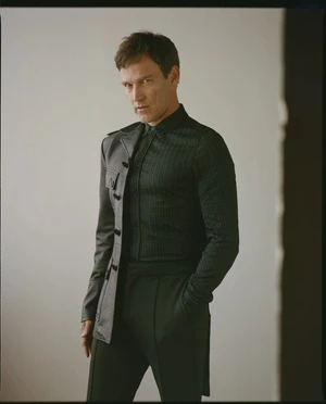 Man wearing a dark patterned button-down shirt and a half-leather jacket standing against a plain background