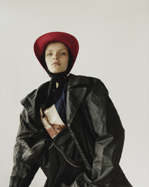 Model wearing a red brimmed hat and dark trench coat against a plain white studio background.
