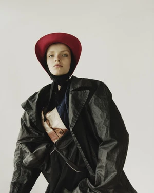 Model wearing a red brimmed hat and dark trench coat against a plain white studio background.