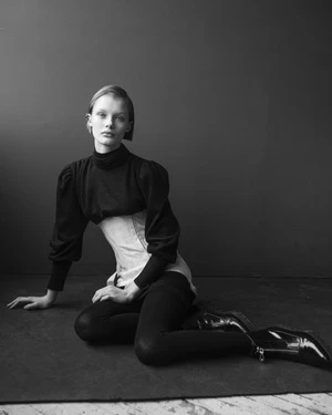 A young fair-skinned woman in a black turtleneck, corset, and leggings sits on the floor in a high-contrast black-and-white portrait.