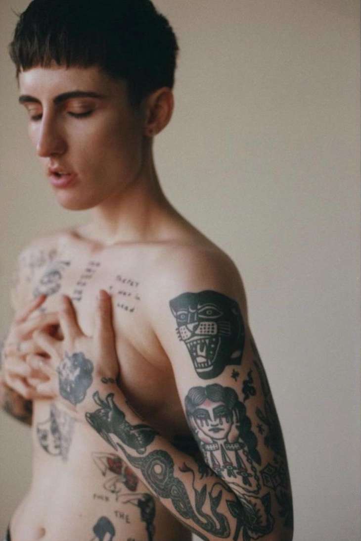 Shirtless person with tattoos covering torso and arms, eyes closed, standing against a plain background in an indoor setting.