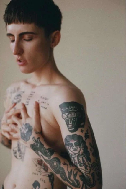 Shirtless person with tattoos covering torso and arms, eyes closed, standing against a plain background in an indoor setting.
