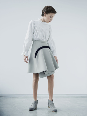 A young person in a stylish white blouse and asymmetrical skirt poses in a brightly lit studio with a smooth, light gray floor.