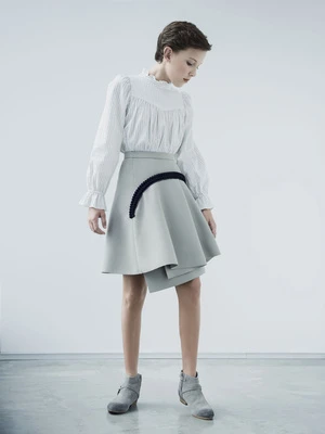 A young person in a stylish white blouse and asymmetrical skirt poses in a brightly lit studio with a smooth, light gray floor.