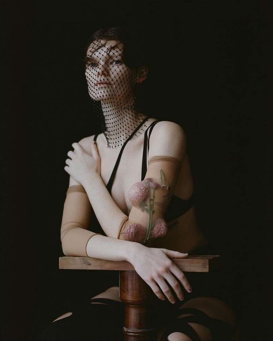 A fair-skinned woman poses on a wooden stool, wearing a black veil and bra, sheer arm coverings, and striped thigh-highs against a dark backdrop.