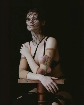 A fair-skinned woman poses on a wooden stool, wearing a black veil and bra, sheer arm coverings, and striped thigh-highs against a dark backdrop.