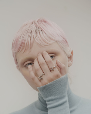 Close-up studio portrait of a fair-skinned adult woman with pastel pink hair, wearing a blue turtleneck and silver rings on her hand.