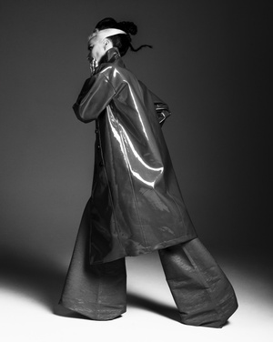 Black and white studio portrait of a model wearing a shiny coat and wide-leg pants, looking over their shoulder with a hand near their face.