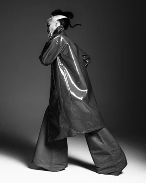 Black and white studio portrait of a model wearing a shiny coat and wide-leg pants, looking over their shoulder with a hand near their face.