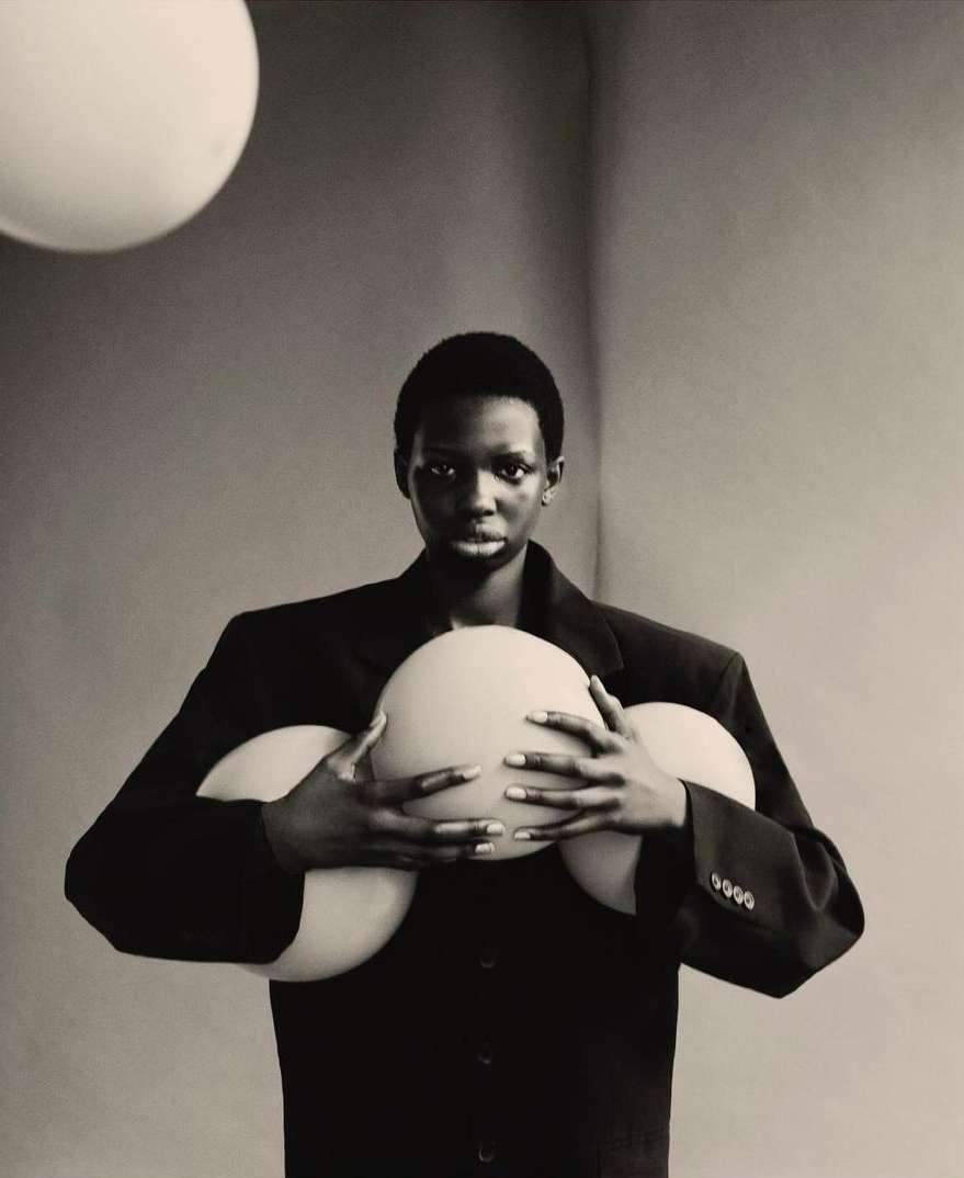 Black man holding three large spherical orbs while wearing an oversized dark suit jacket against a plain grey studio background.