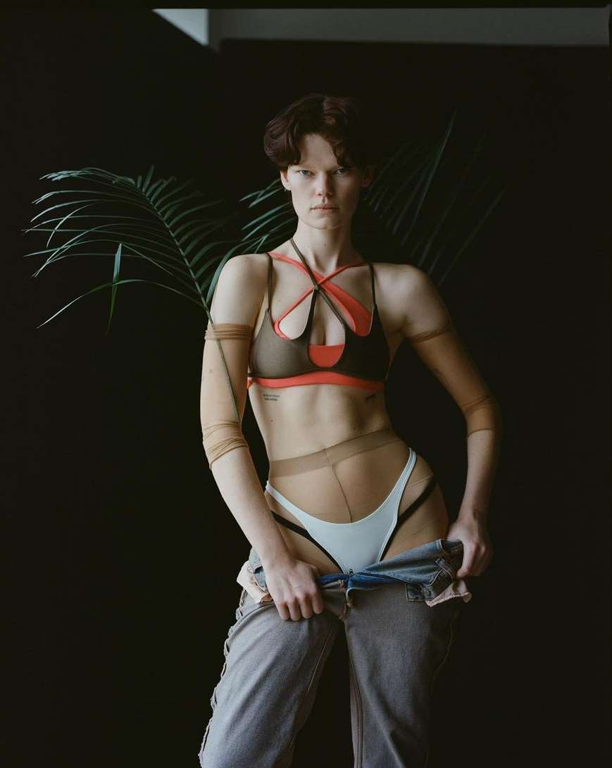 Stylish woman poses in bra, pants, and hose in a photo studio with black backdrop and a green plant, fashion photoshoot, medium shot.