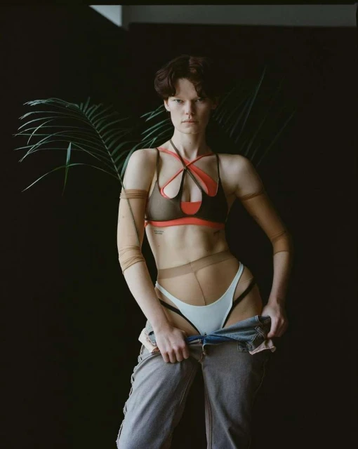 Stylish woman poses in bra, pants, and hose in a photo studio with black backdrop and a green plant, fashion photoshoot, medium shot.