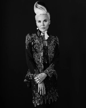 Portrait of a blonde woman with dark hair underneath, wearing an embroidered top and pearl earrings, set against a black backdrop.