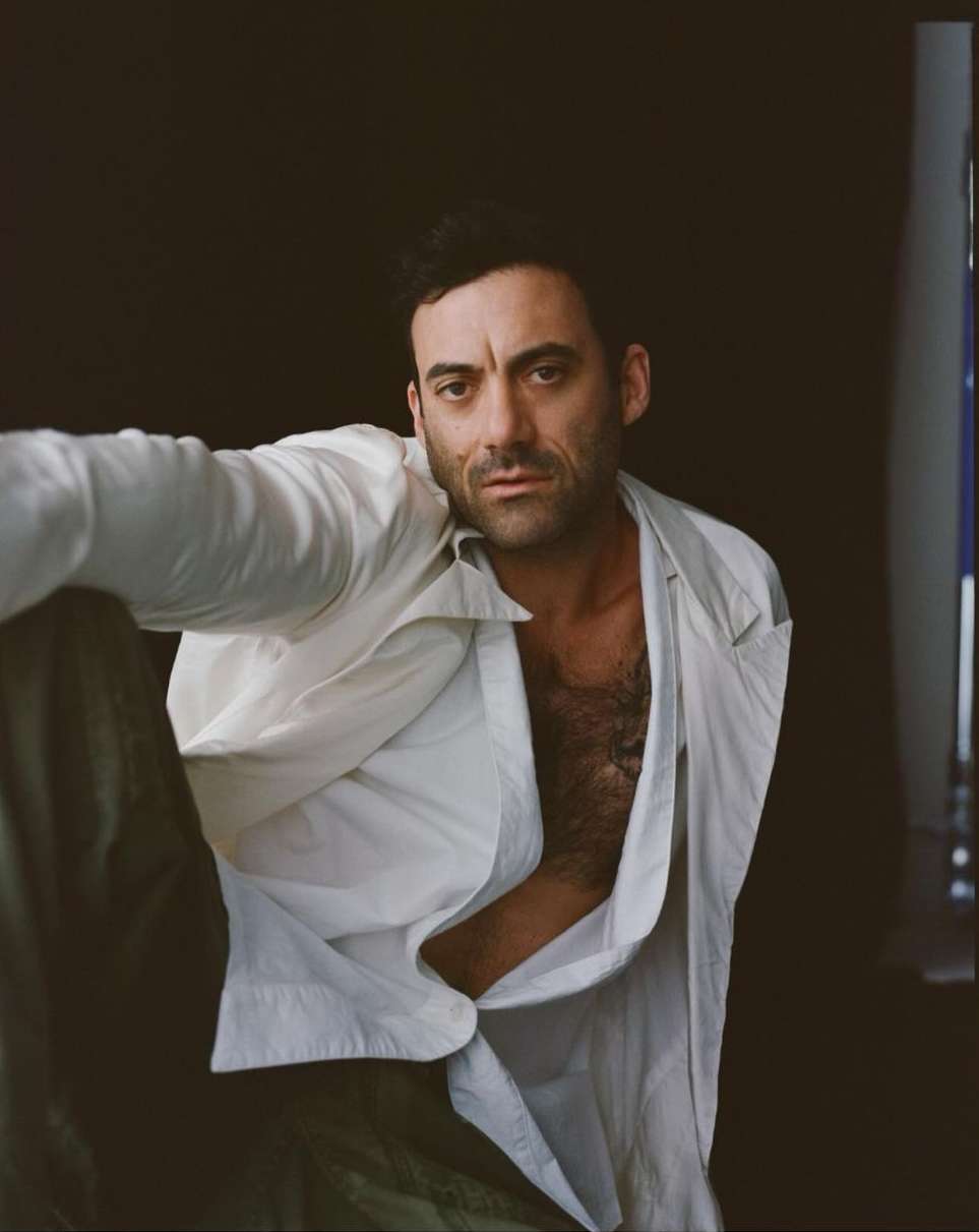 Portrait of adult male in a partially unbuttoned white shirt against a dark backdrop, looking at the camera with a neutral expression.