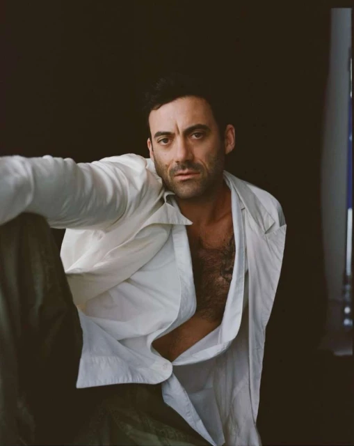 Portrait of adult male in a partially unbuttoned white shirt against a dark backdrop, looking at the camera with a neutral expression.