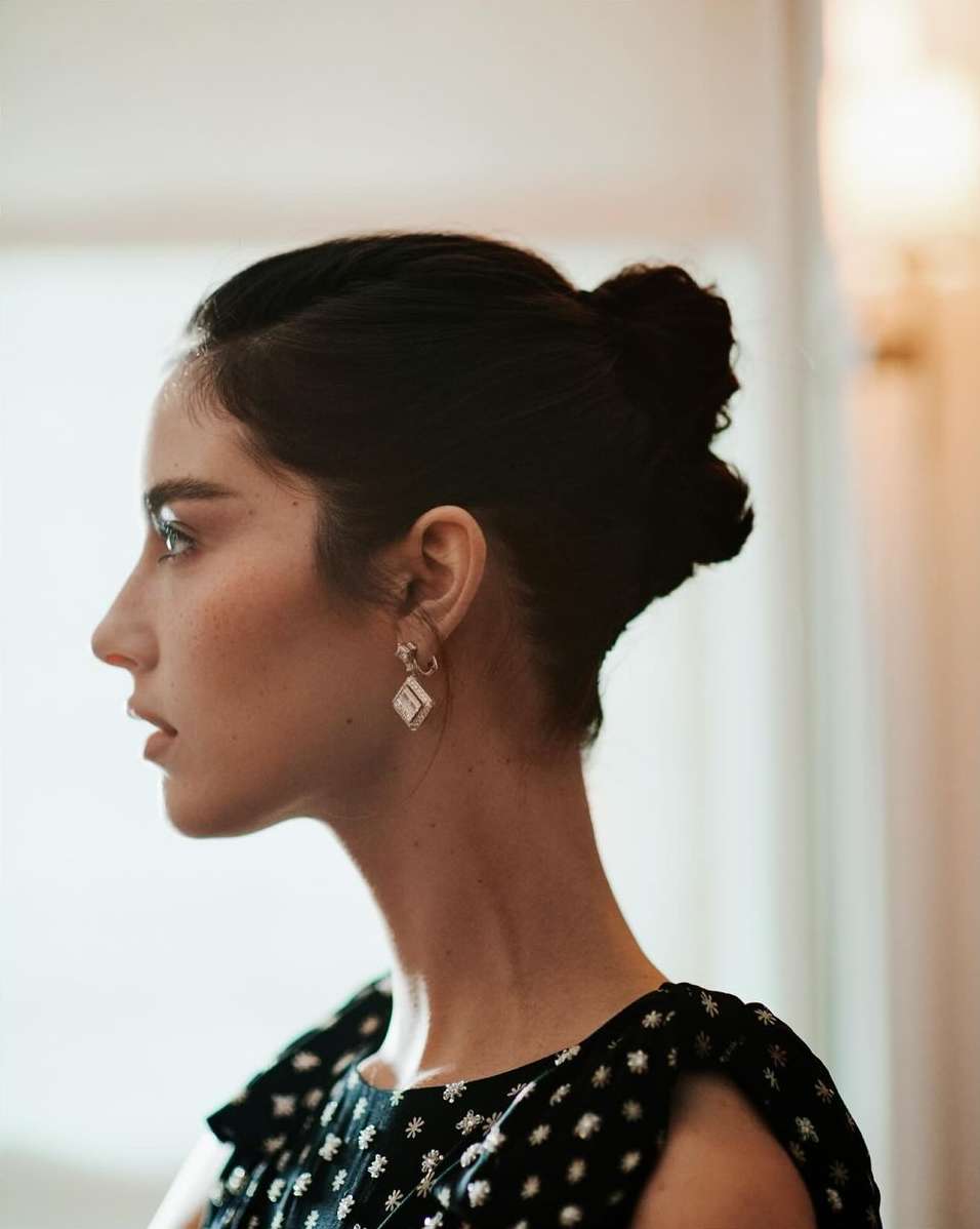 Profile portrait of a woman with dark hair in a bun wearing patterned dark top and diamond earrings by Matthew Priestley