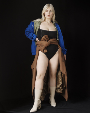 Full-length studio portrait of a plus-size female model in a black bodysuit with a brown wrap, blue jacket and cream boots on black background.