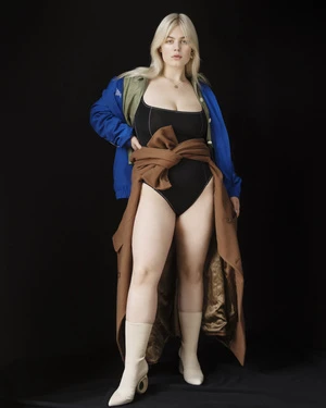 Full-length studio portrait of a plus-size female model in a black bodysuit with a brown wrap, blue jacket and cream boots on black background.