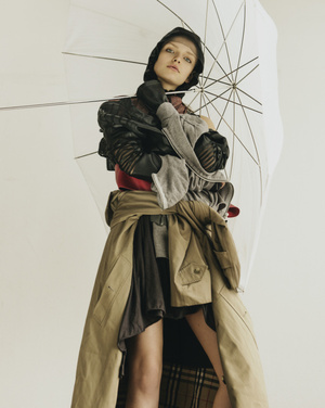 Fashion shot of a woman wearing layered garments and an umbrella, photographed indoors against a plain backdrop in soft, diffused lighting.