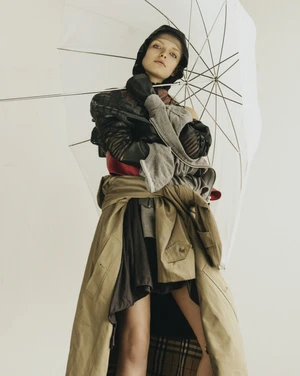 Fashion shot of a woman wearing layered garments and an umbrella, photographed indoors against a plain backdrop in soft, diffused lighting.