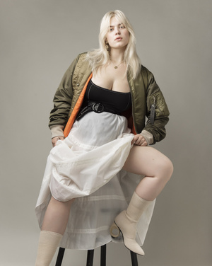 Woman with blonde hair poses seated wearing a bomber jacket, black top, white skirt, and boots in a studio against a gray background.