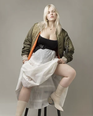 Woman with blonde hair poses seated wearing a bomber jacket, black top, white skirt, and boots in a studio against a gray background.