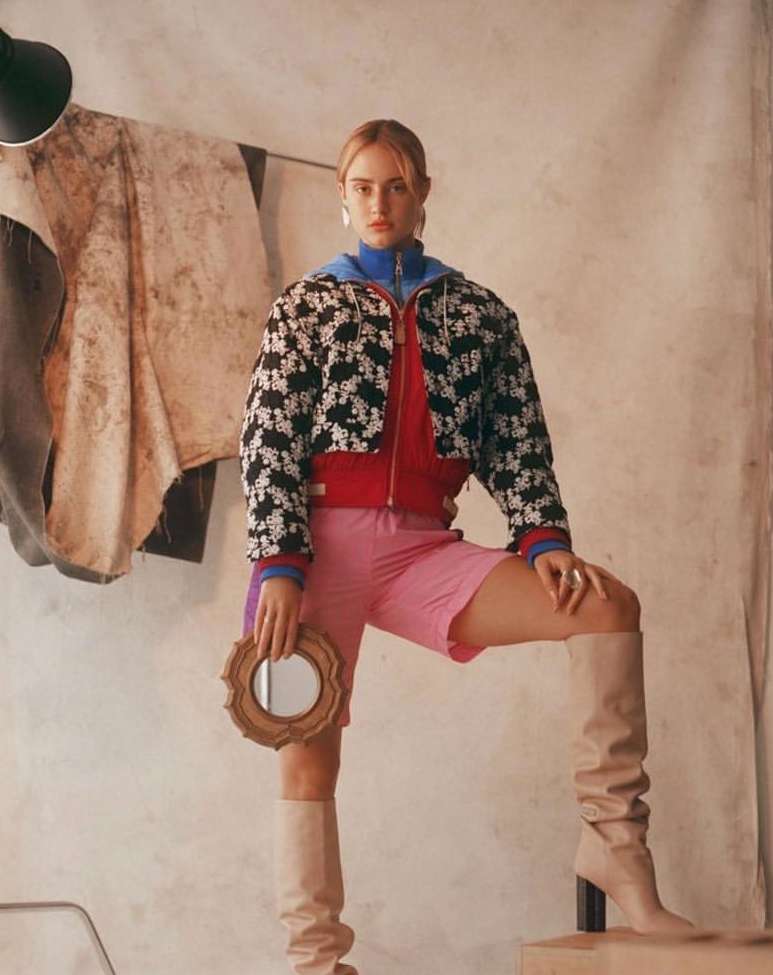 Fashionable woman in jacket, shorts and knee-high boots poses indoors, holding a framed mirror. Neutral tones and soft lighting.