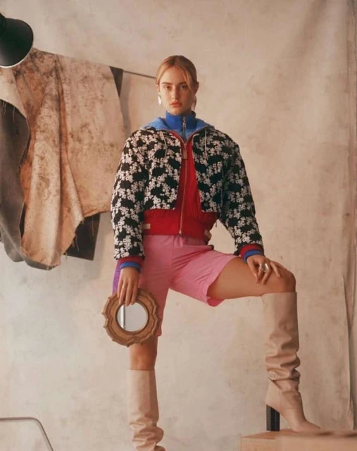 Fashionable woman in jacket, shorts and knee-high boots poses indoors, holding a framed mirror. Neutral tones and soft lighting.