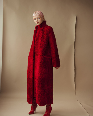 Woman with pink hair models a striking full-length red textured coat with matching high-heeled boots against a beige backdrop.