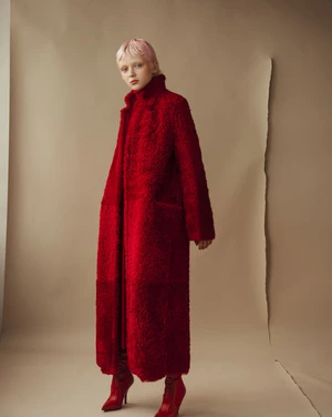 Woman with pink hair models a striking full-length red textured coat with matching high-heeled boots against a beige backdrop.