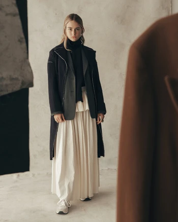 Woman in a dark coat and cream pleated skirt standing before a textured grey wall
