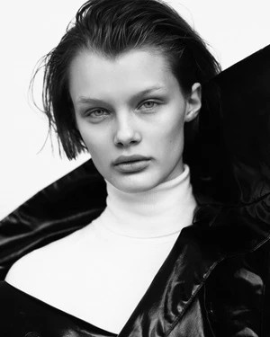 A close-up portrait of a young adult female model with short dark hair, wearing a white turtleneck and a dark vinyl coat in a studio setting.