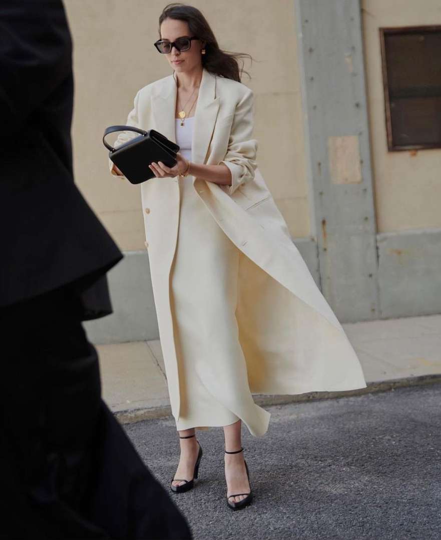 Woman in a cream long coat holding a black handbag walking down an urban street