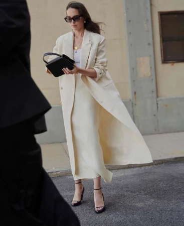 Woman in a cream long coat holding a black handbag walking down an urban street