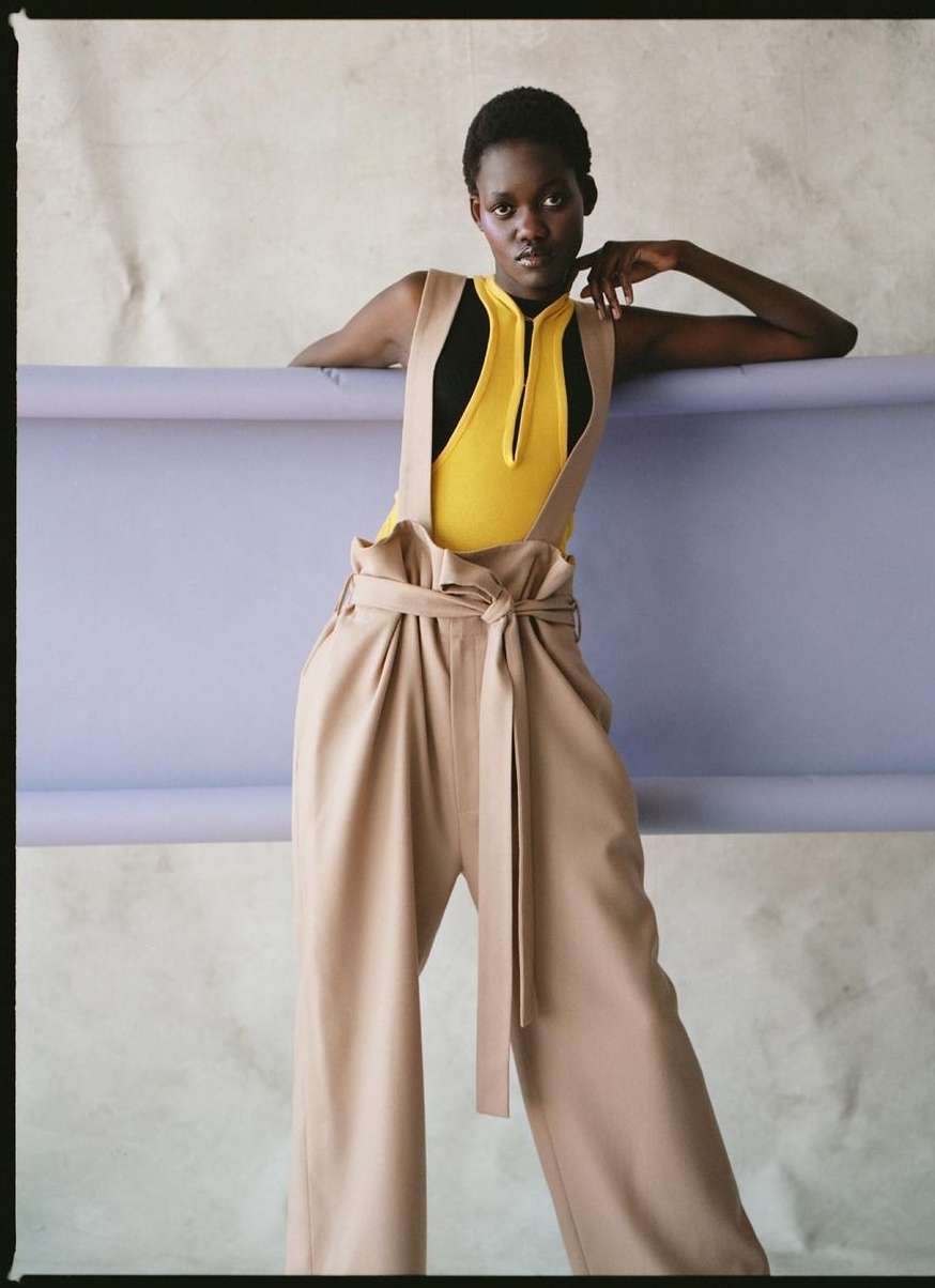 Fashionable woman in wide-leg pants and bodysuit strikes a pose against a lilac and white studio backdrop, showcasing modern style.