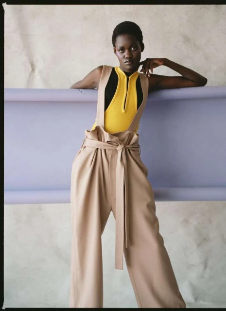 Fashionable woman in wide-leg pants and bodysuit strikes a pose against a lilac and white studio backdrop, showcasing modern style.