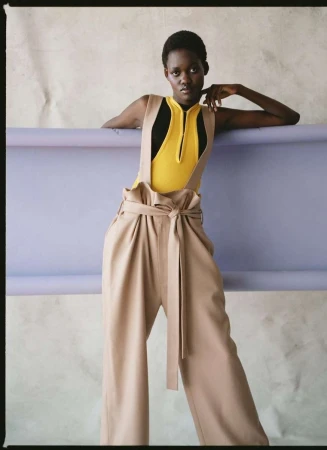 Fashionable woman in wide-leg pants and bodysuit strikes a pose against a lilac and white studio backdrop, showcasing modern style.