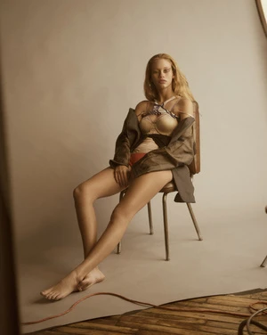 Young woman poses barefoot on chair in photo studio, wearing jacket and patterned lingerie, captured in medium shot with soft lighting.