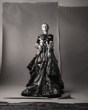 Full-length shot of a fair-skinned fashion model in an elaborate floral gown with a large bow, posing against a gray backdrop.